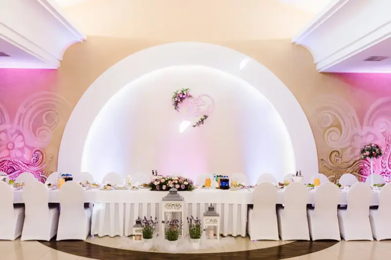 Elegancka sala weselna z białymi pokrowcami na krzesłach i serwowanym stołem. Serce z imionami pary młodej zdobi ścianę, tworząc atmosferę wesela z klasą.